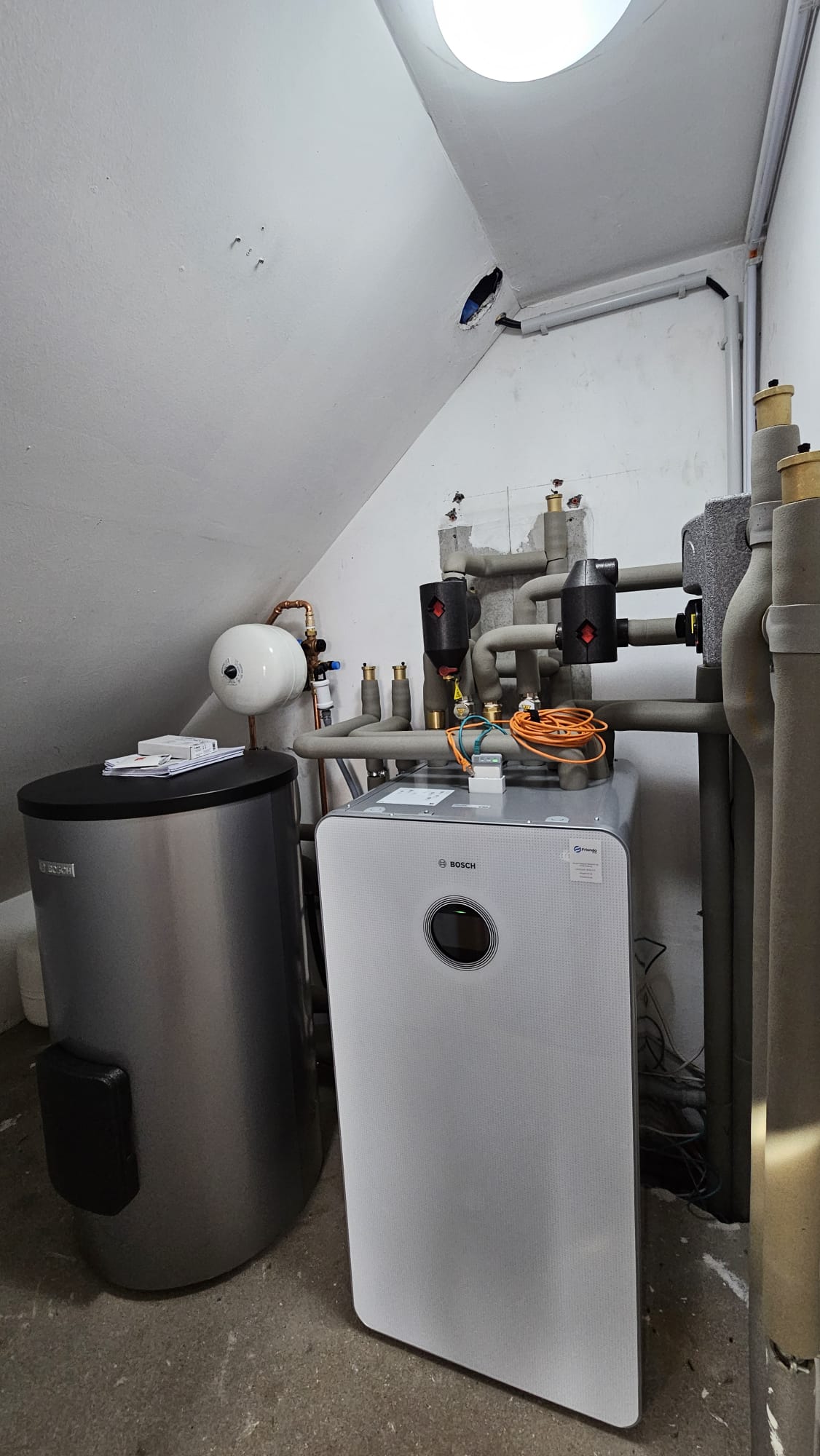 Eine Bosch-Wärmepumpen-Inneneinheit steht kompakt im Heizungskeller, angeschlossen an Speicher und umfangreiche Rohrleitungen.